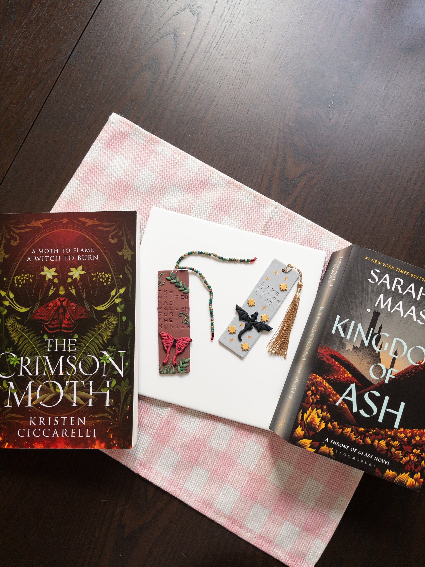 Two books, 'The Crimson Moth' and 'Kingdom of Ash', on a checkered surface with decorative bookmarks.