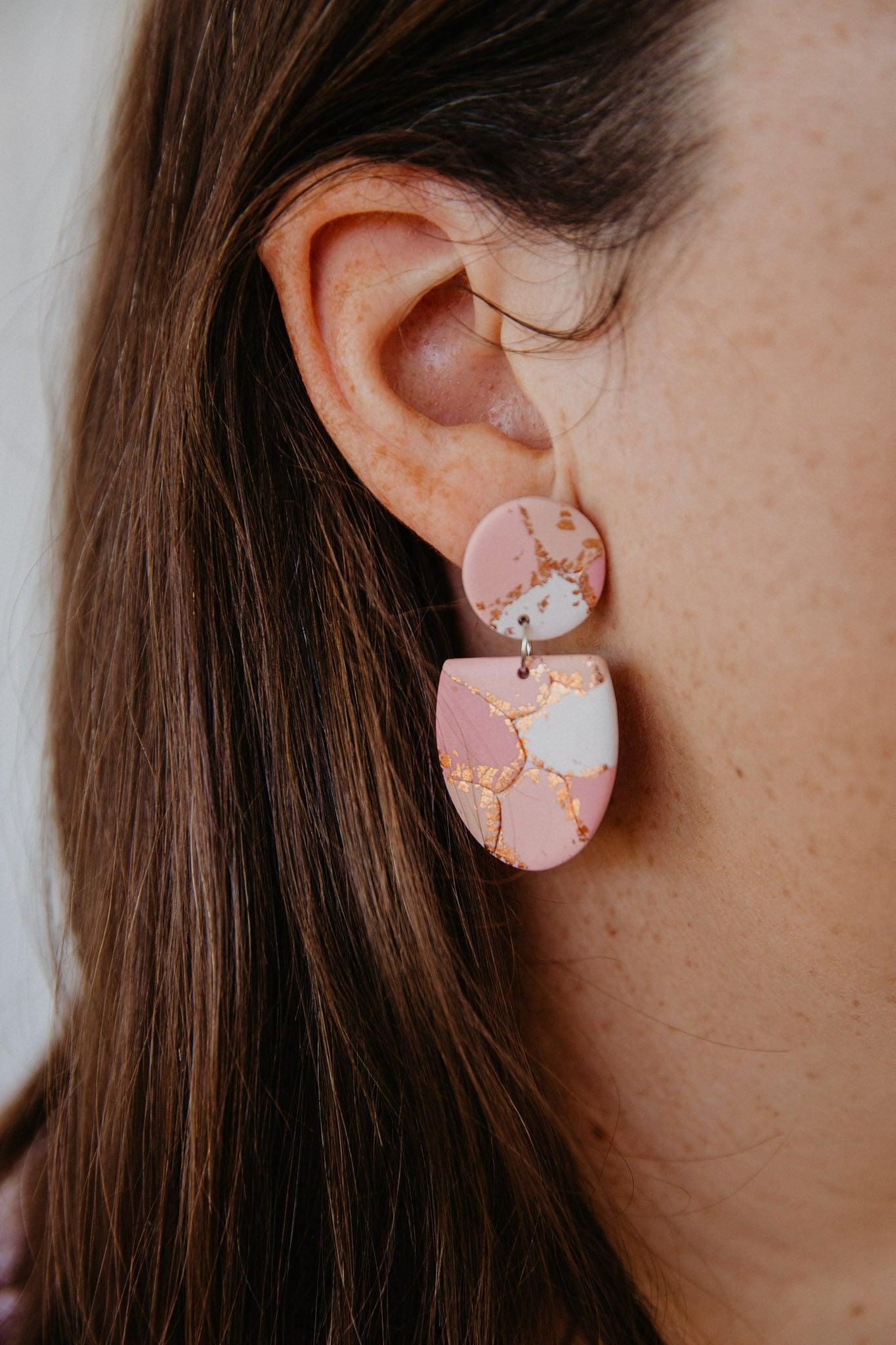 Close up on woman's ear wearing a half oval dangle with a circle stud top. Dangles feature a pattern of patches of maroon, orange and cream with copper foil between.