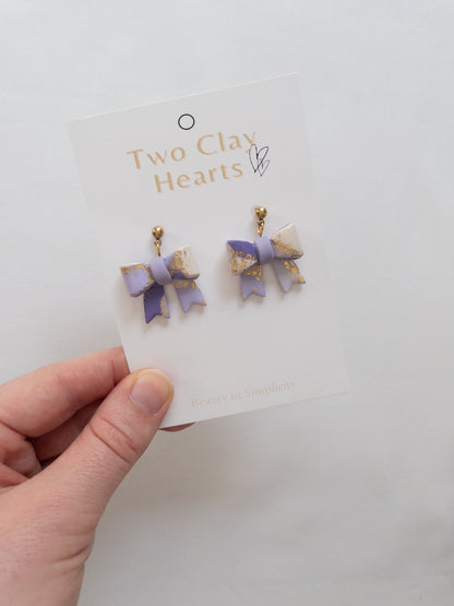 Pair of bow-shaped earrings on a 'Two Clay Hearts' branded card held by a hand.
