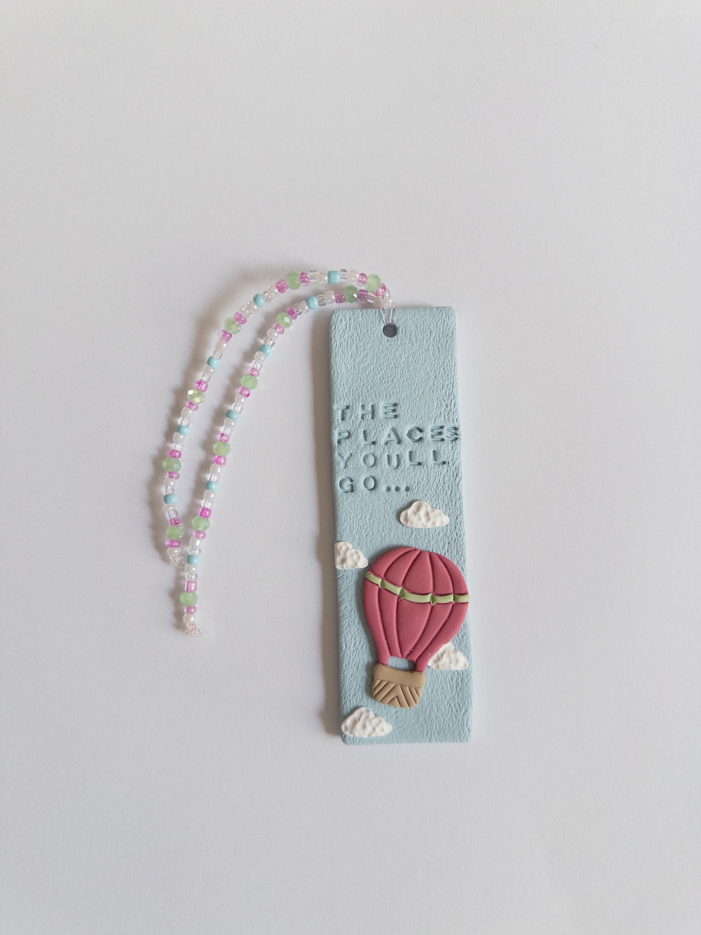 Polymer clay bookmark and matching beaded tassel on white background. Bookmark features words 'the places you'll go' on a blue background, with white clouds and a pink hot air balloon.