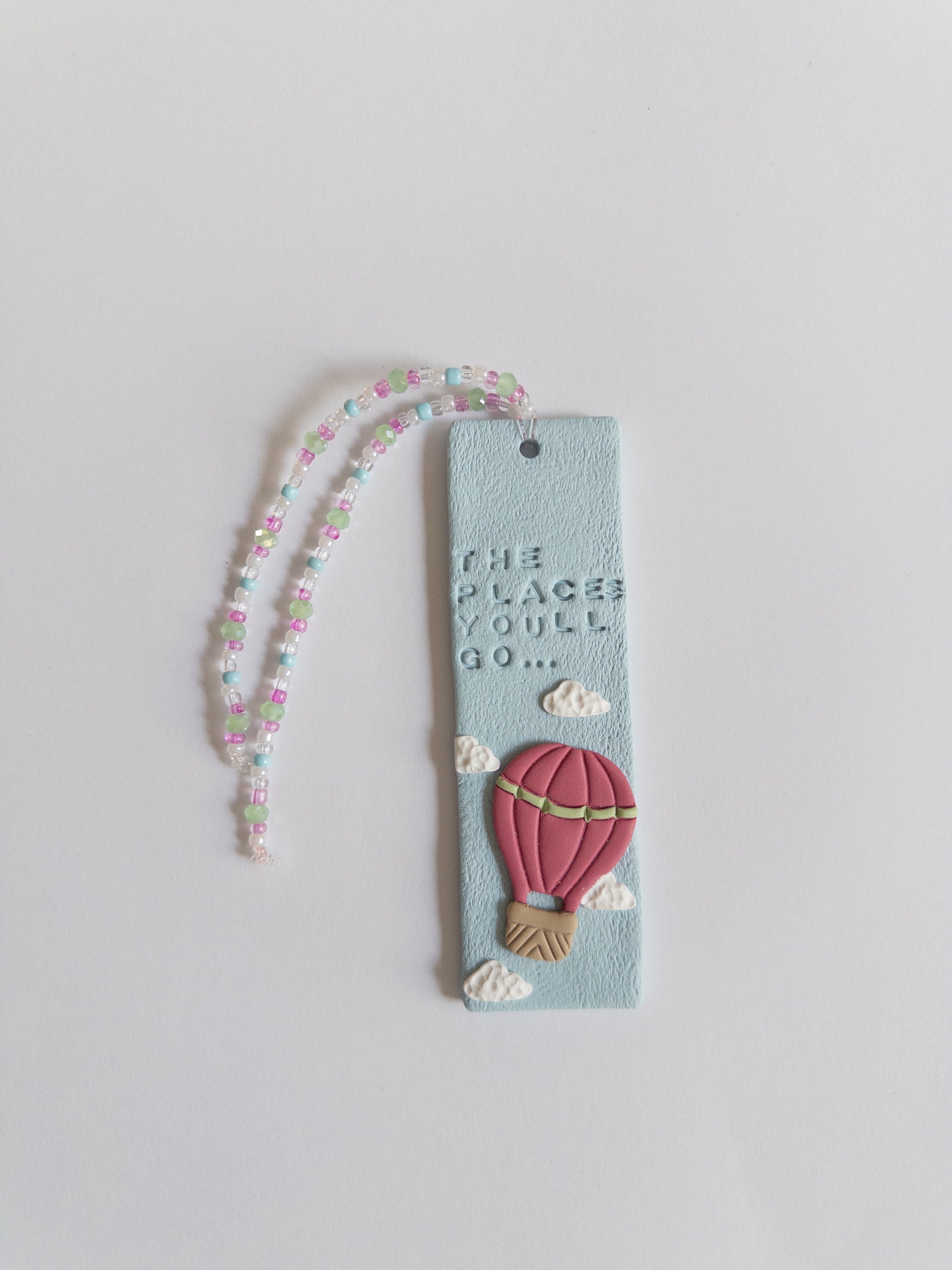 Polymer clay bookmark and matching beaded tassel on white background. Bookmark features words 'the places you'll go' on a blue background, with white clouds and a pink hot air balloon.