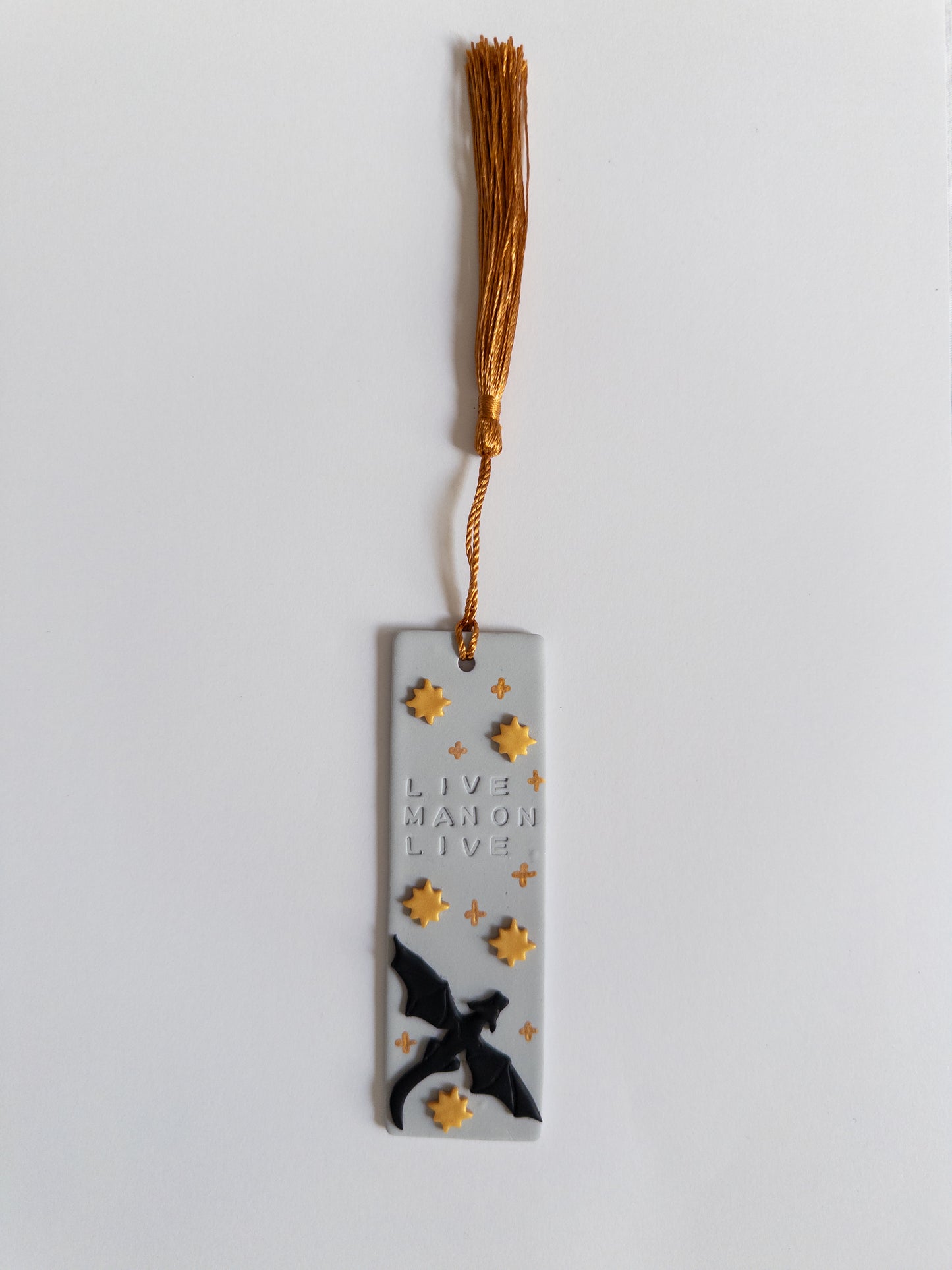 Grey polymer clay bookmark with gold coloured tassel on white background. Bookmark features a dragon, gold stars and the words 'live Manon live'