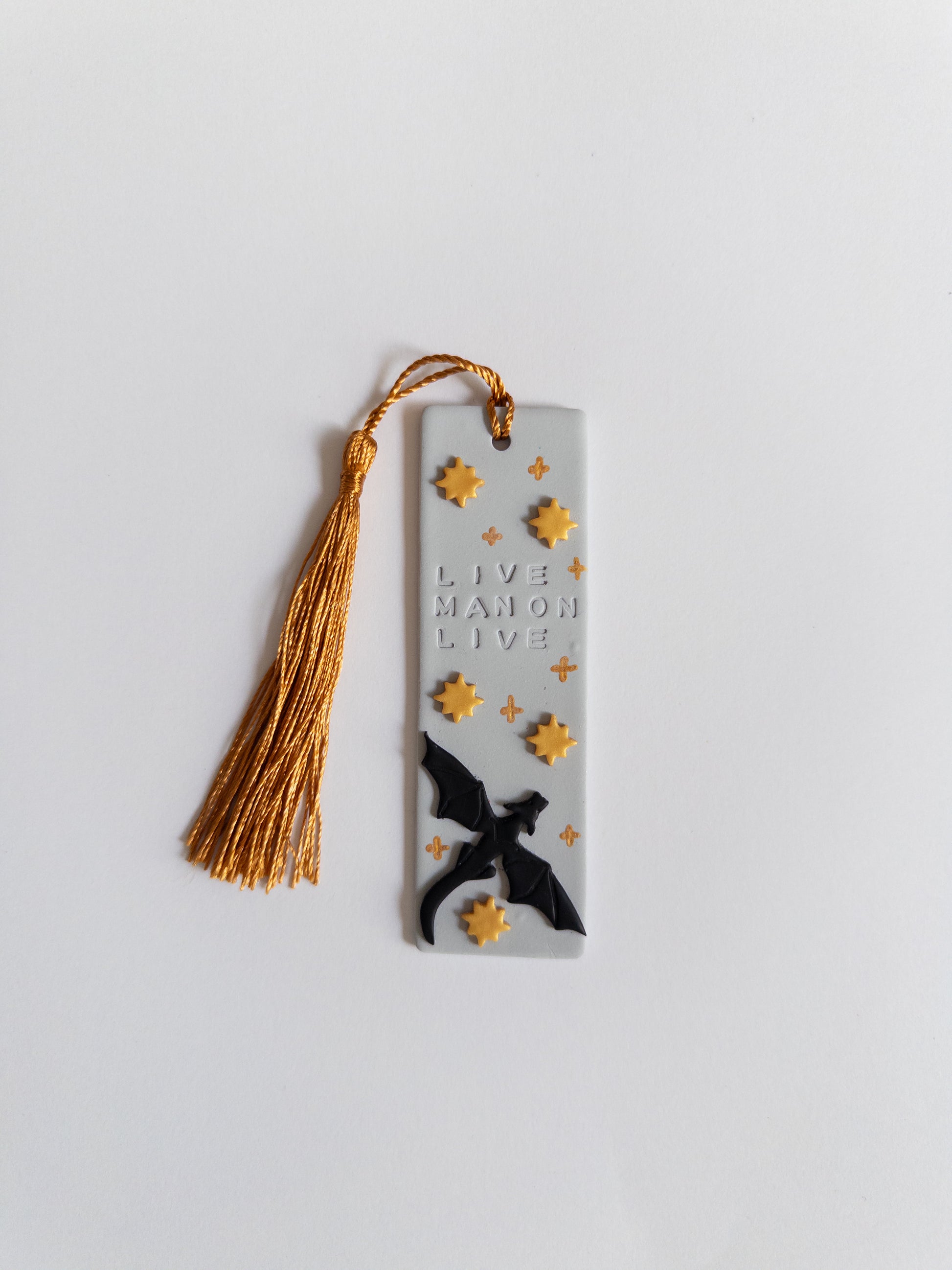 Grey polymer clay bookmark with gold coloured tassel on white background. Bookmark features a dragon, gold stars and the words 'live Manon live'