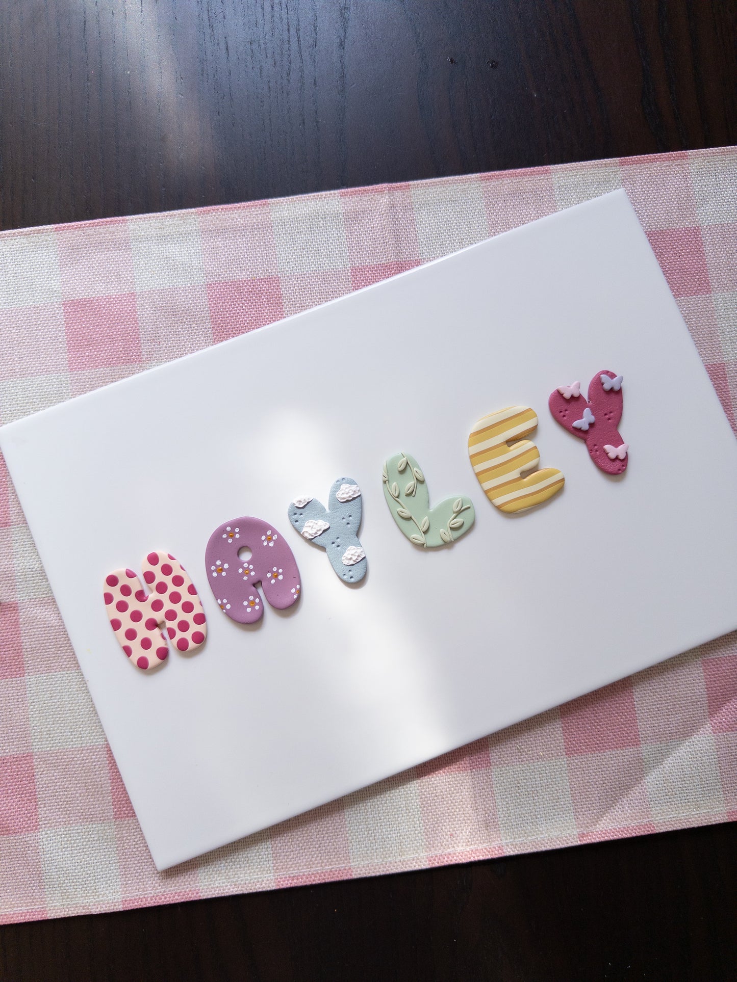 A white rectangular card placed on a pink-and-white checkered mat, with the word "Hayley" spelled out by colorful decorative letters on the surface.