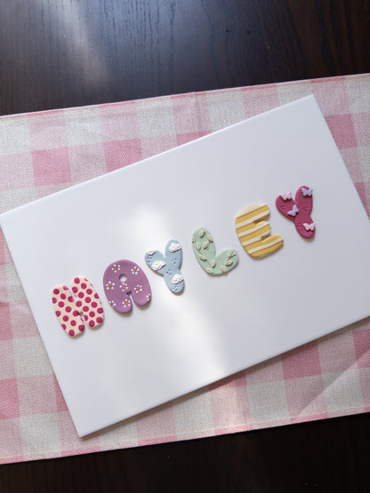 A white rectangular card placed on a pink-and-white checkered mat, with the word "Hayley" spelled out by colorful decorative letters on the surface.