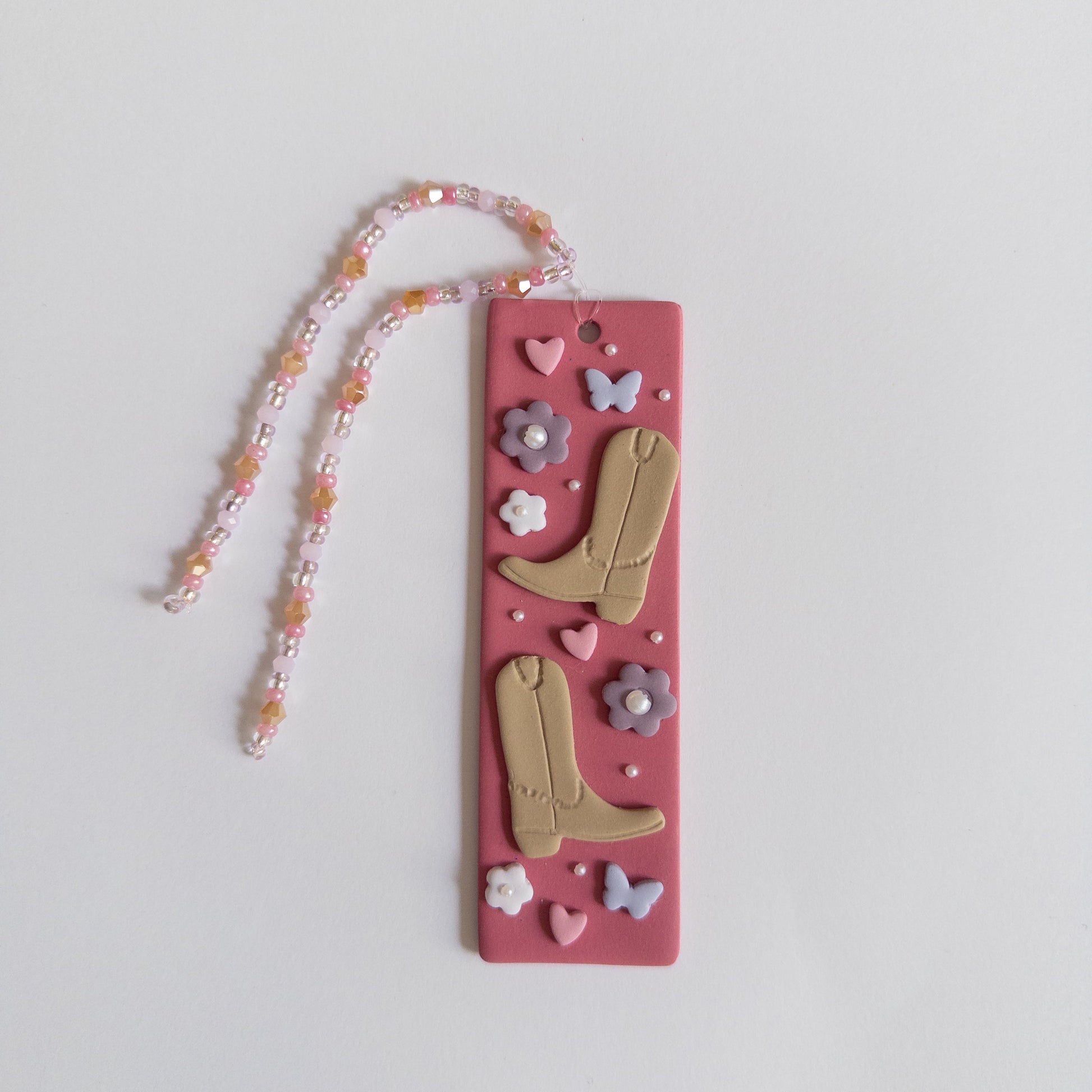 Polymer clay bookmark with a pink background and decorated with brown cowboy boots, purple flowers and butterflies and pink hearts and a matching beaded tassel