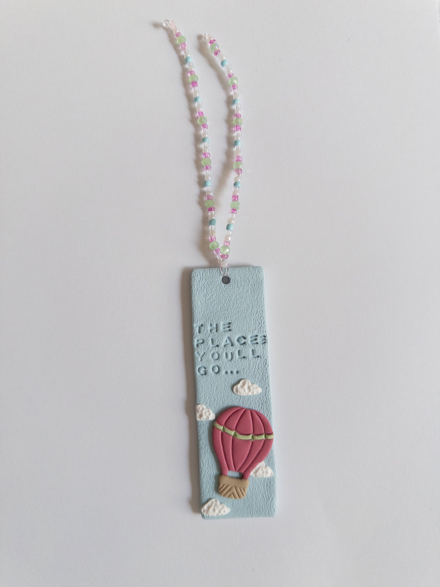 Polymer clay bookmark and matching beaded tassel on white background. Bookmark features words 'the places you'll go' on a blue background, with white clouds and a pink hot air balloon.