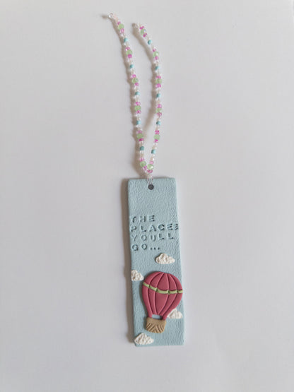 Polymer clay bookmark and matching beaded tassel on white background. Bookmark features words 'the places you'll go' on a blue background, with white clouds and a pink hot air balloon.