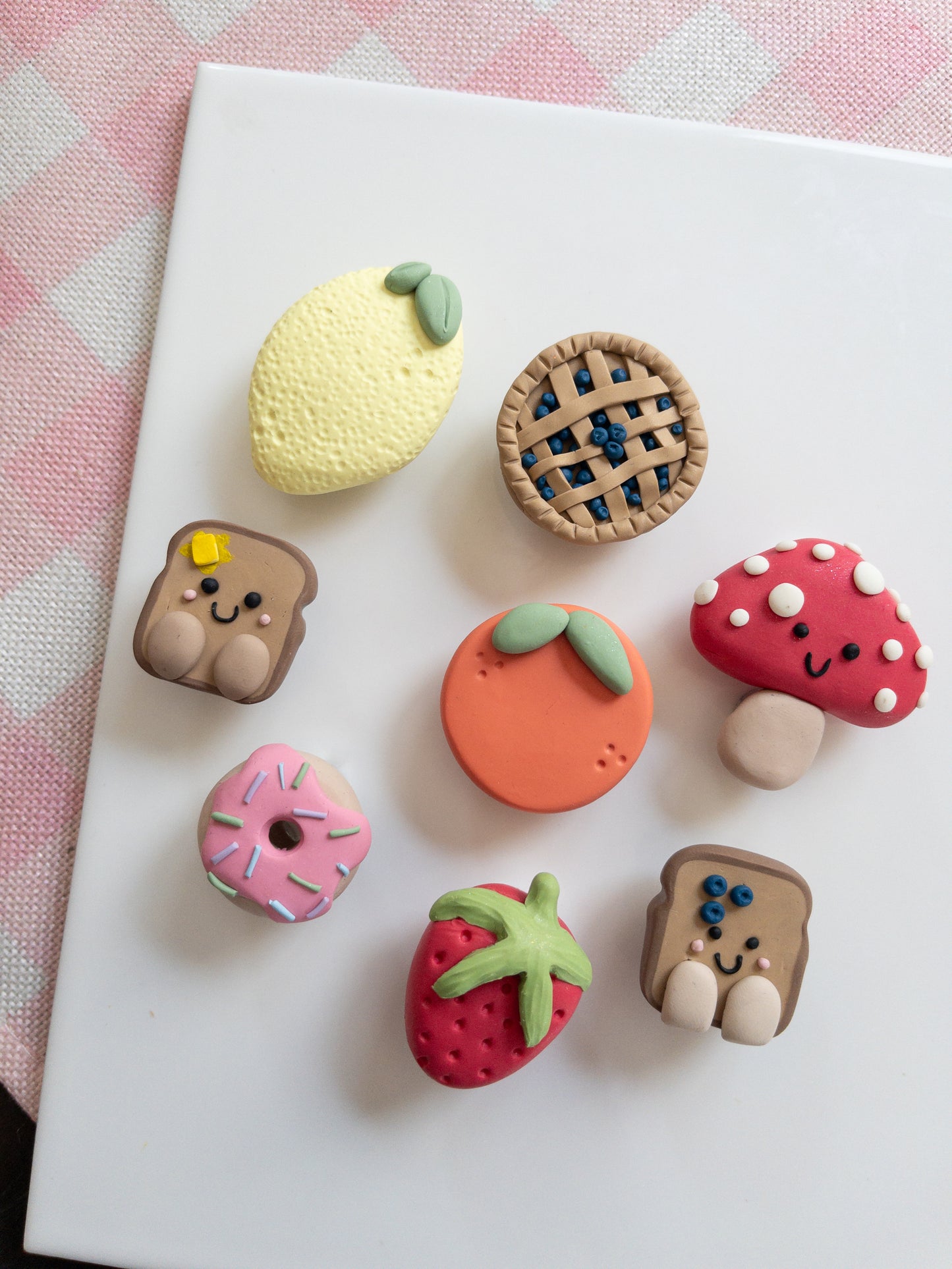 Set of colorful food-themed clay magnets on a white surface with a pink checkered background.