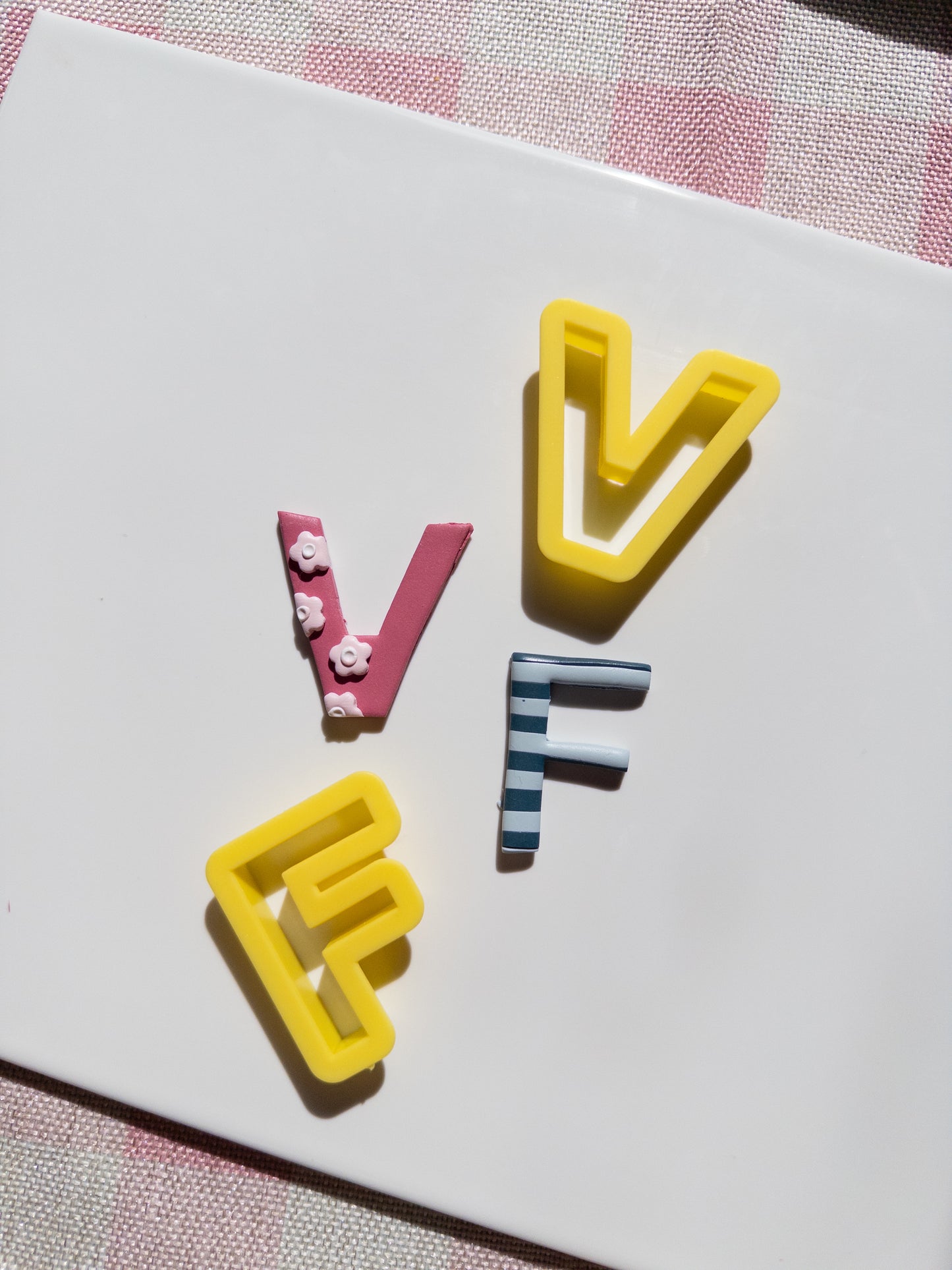 Three decorative clay letters spelling 'V F F' arranged on a white rectangular card, with a pink-and-white checkered fabric visible around the edges.