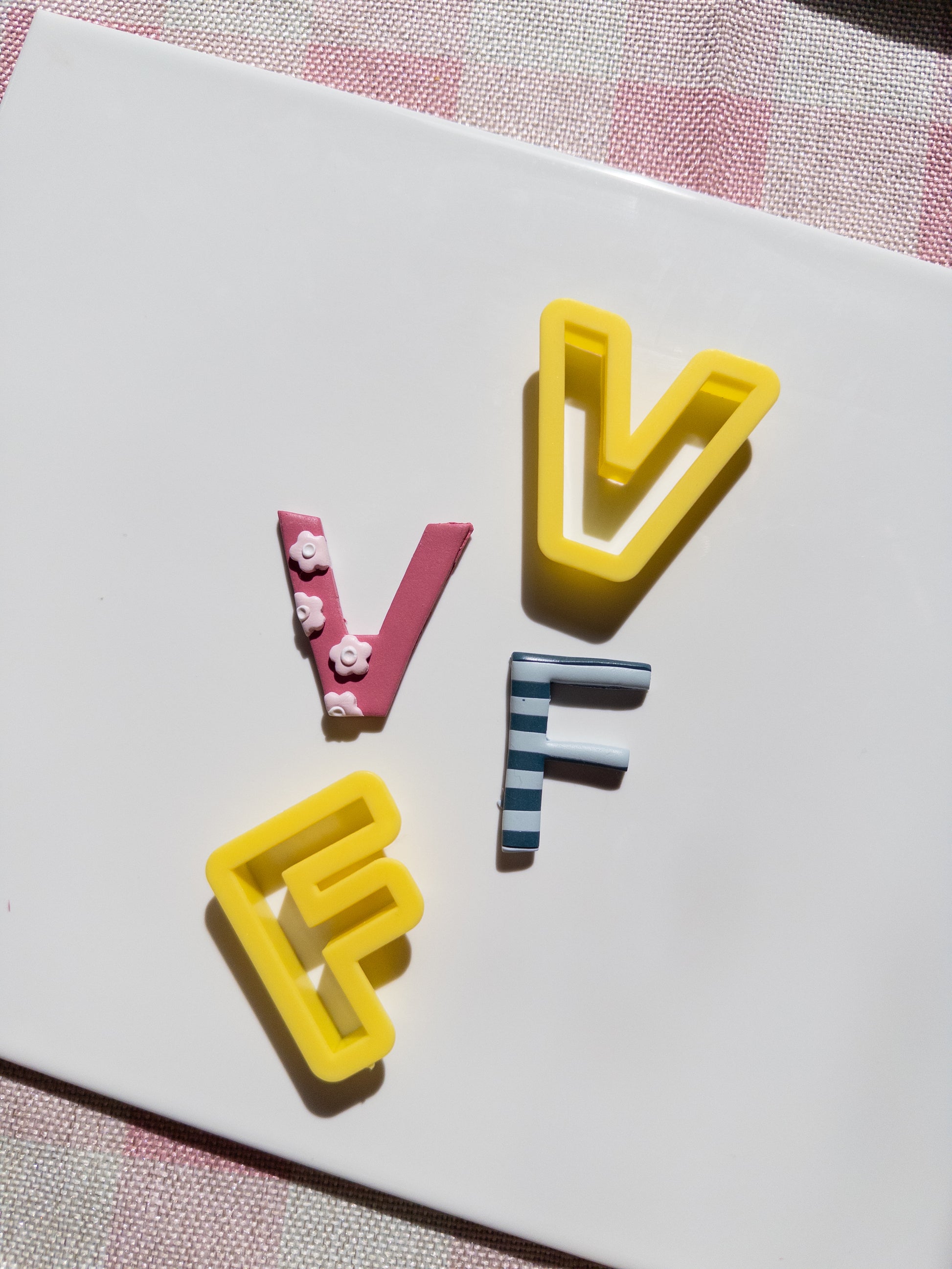 Three decorative clay letters spelling 'V F F' arranged on a white rectangular card, with a pink-and-white checkered fabric visible around the edges.