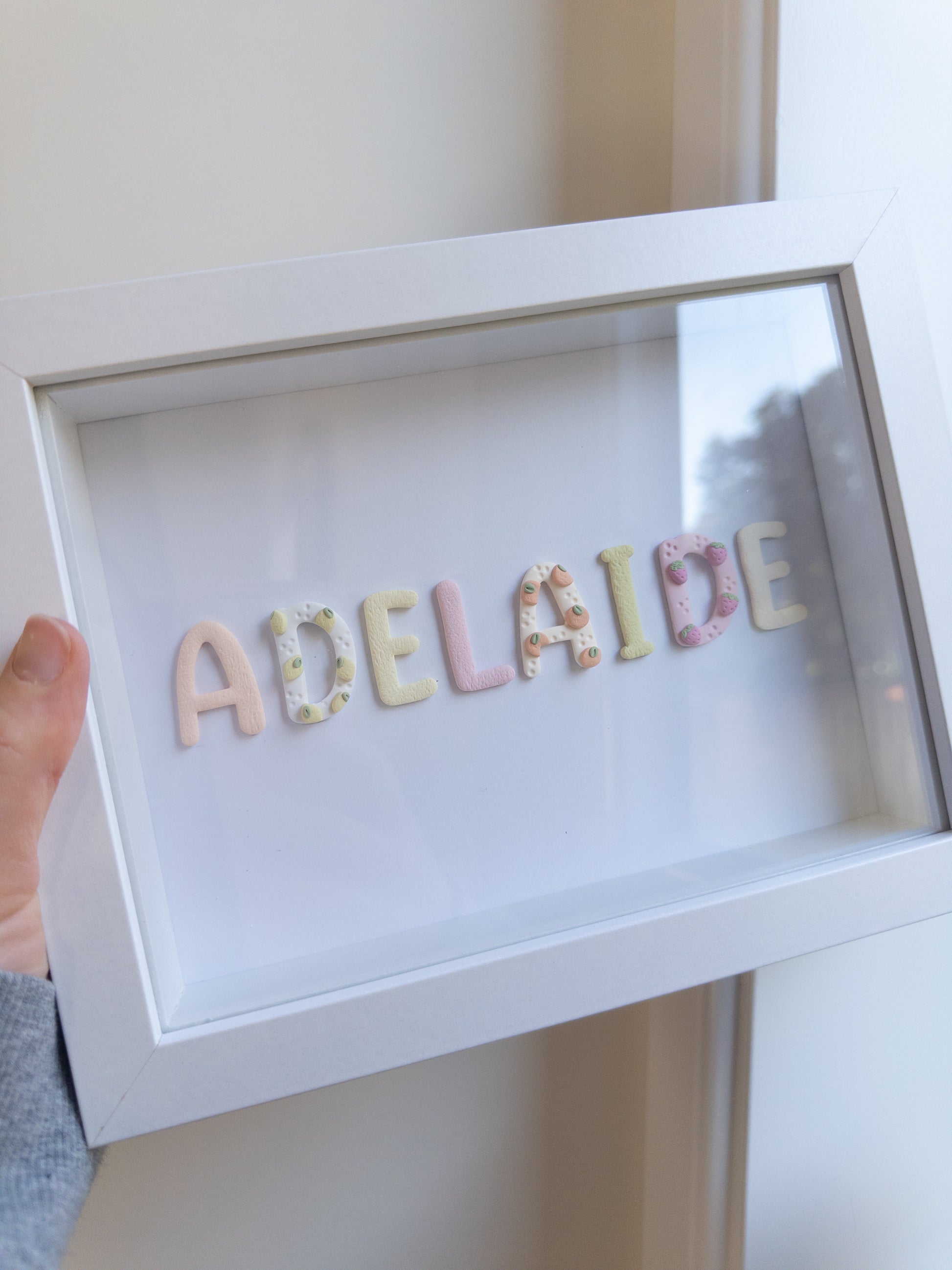 A white shadow box frame with the name 'ADELAIDE' spelled out in pastel decorative letters, held by a hand against a light beige wall.