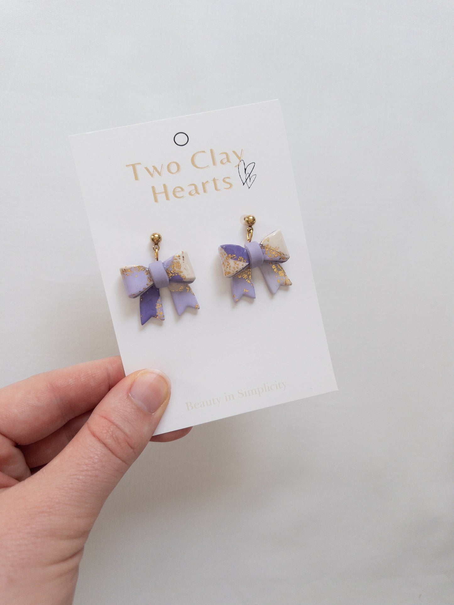 Pair of purple bow-shaped earrings on a 'Two Clay Hearts' branded card held by a hand.