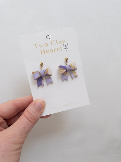 Pair of purple bow-shaped earrings on a 'Two Clay Hearts' branded card held by a hand.