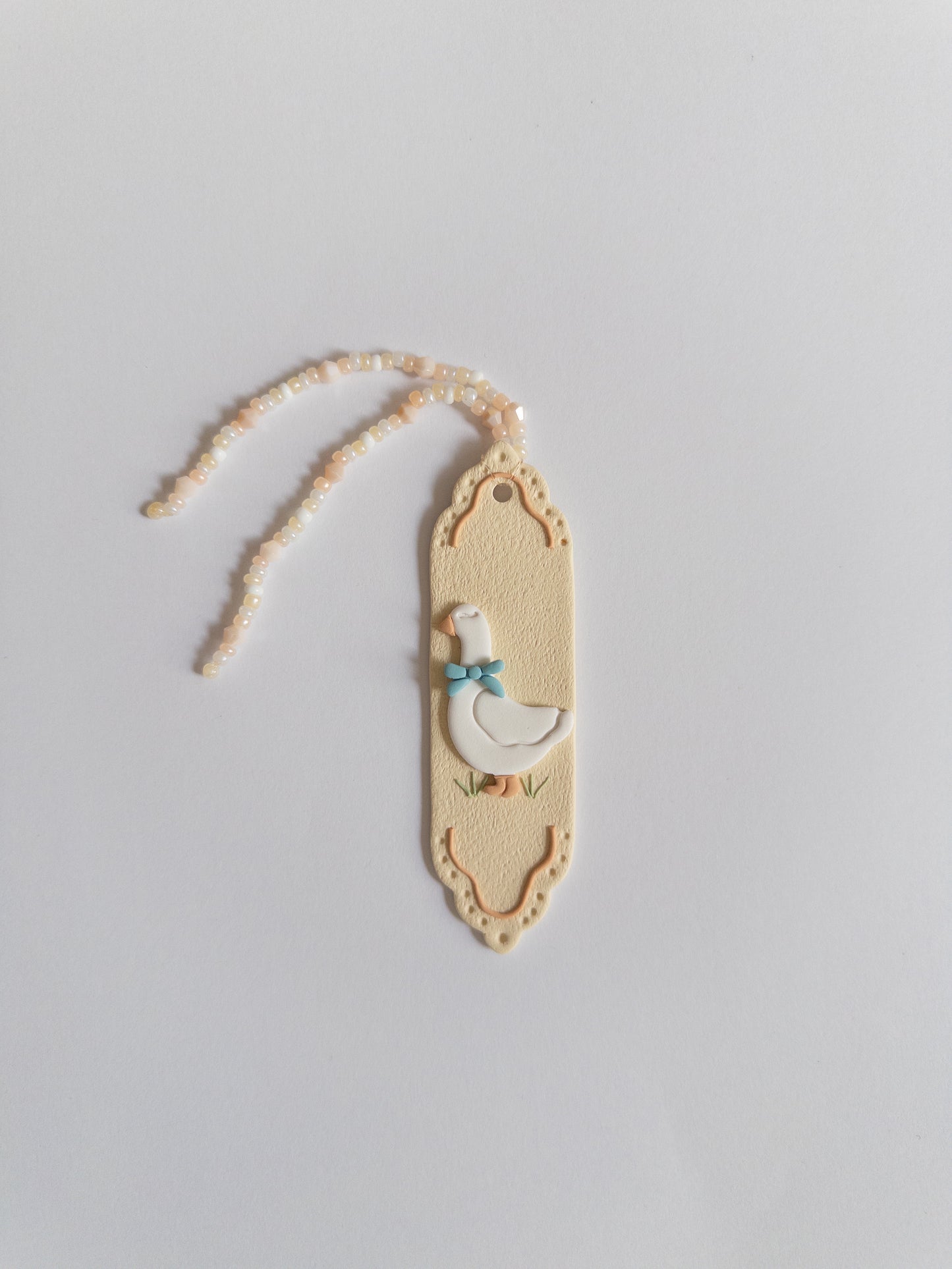 Beige coloured polymer clay bookmark with a white goose on it and a beige and white beaded tassel on a white background