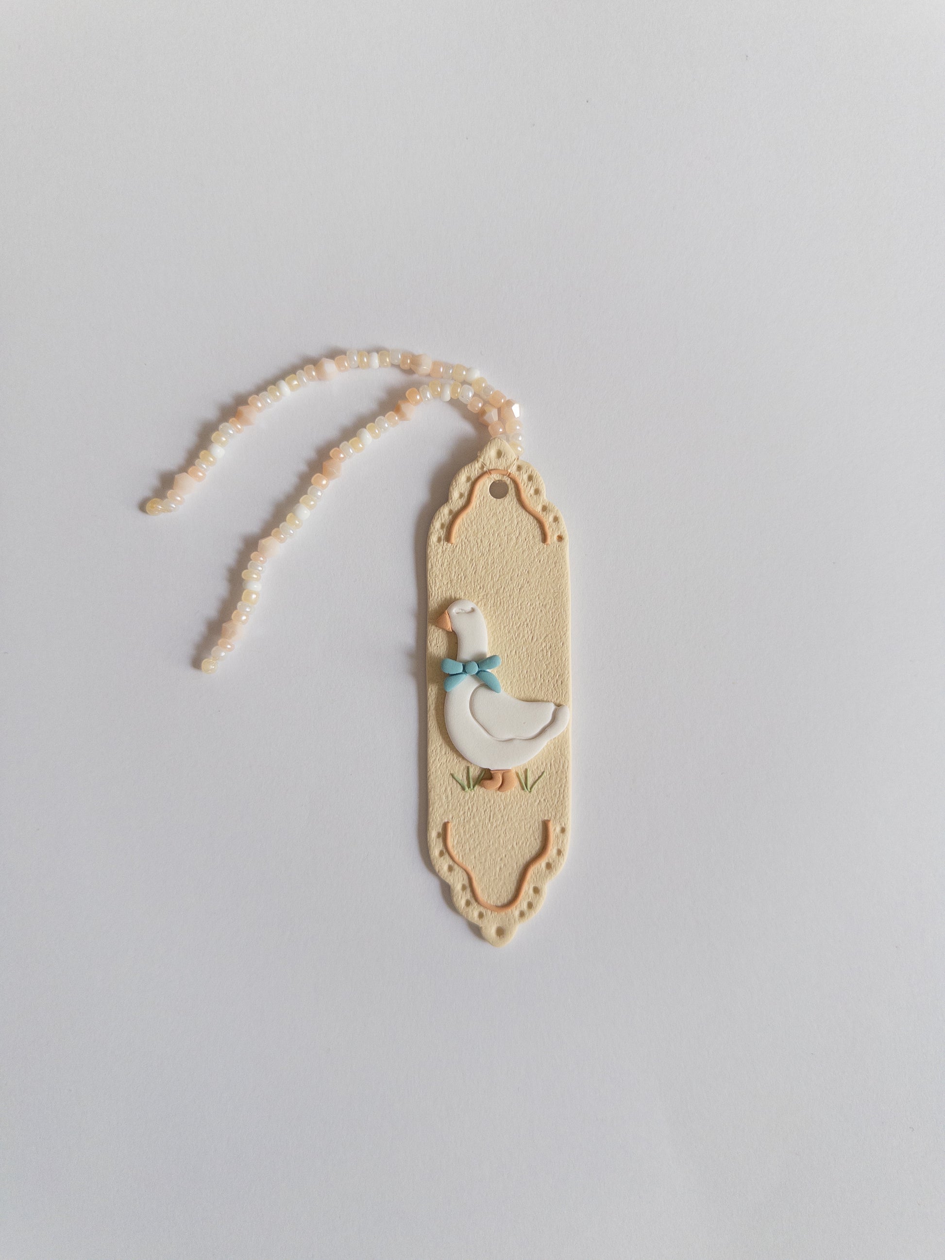 Beige coloured polymer clay bookmark with a white goose on it and a beige and white beaded tassel on a white background