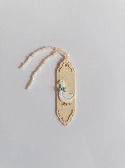 Beige coloured polymer clay bookmark with a white goose on it and a beige and white beaded tassel on a white background