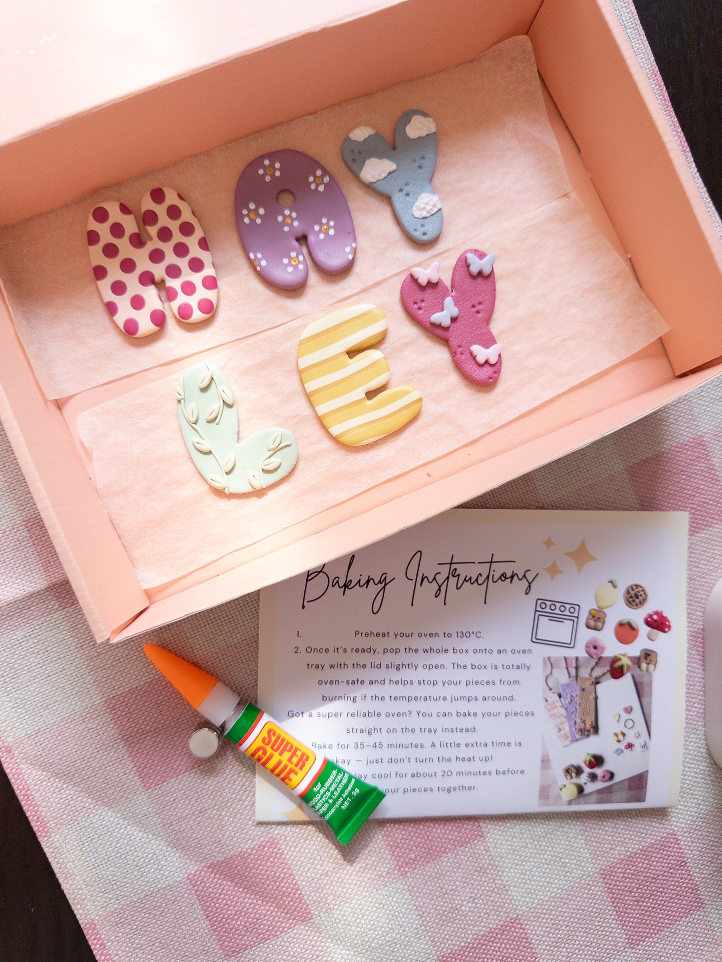 A styled flat-lay of a pink baking box containing colorful clay letter decorations, a small tube of clay glue, and a printed baking instructions card on a pink-and-white checkered cloth.