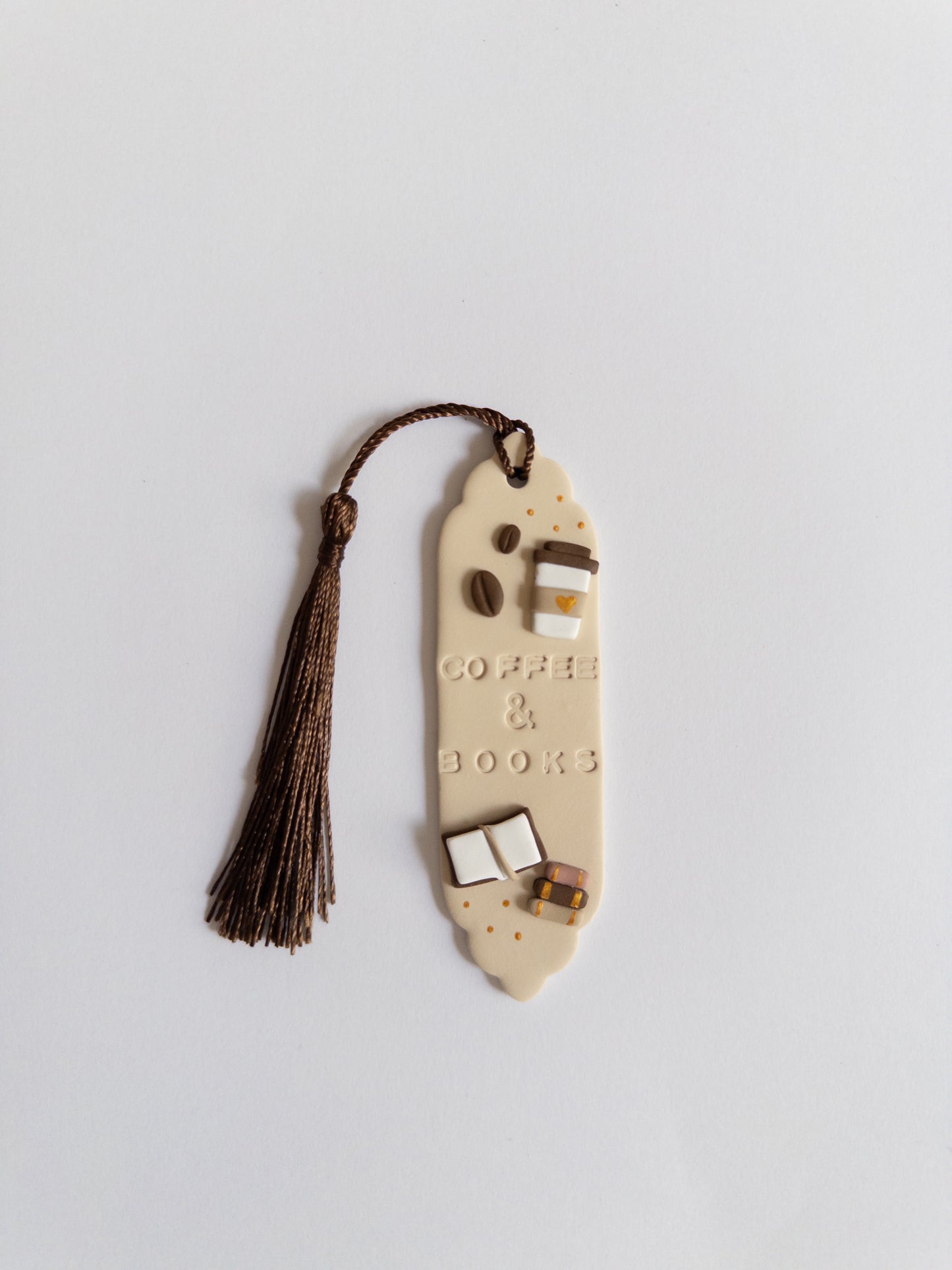 Beige polymer clay bookmark with brown tassel on white background. Bookmark features the words 'coffee & books' and decals of a coffee cup, coffee beans and books.