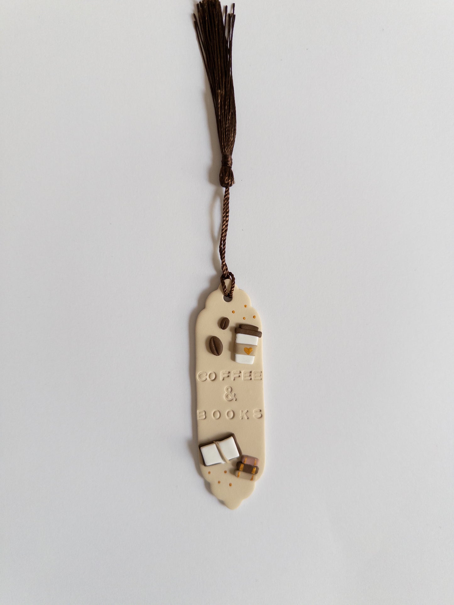 Beige polymer clay bookmark with brown tassel on white background. Bookmark features the words 'coffee & books' and decals of a coffee cup, coffee beans and books.