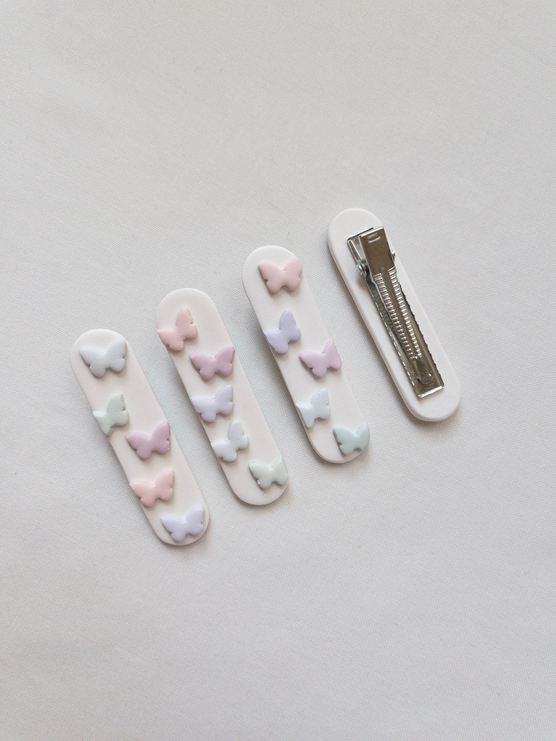 a range of white polymer clay hair clips with pastel butterfly accents on top with one upside down to show the stainless steel alligator hair clip on the back