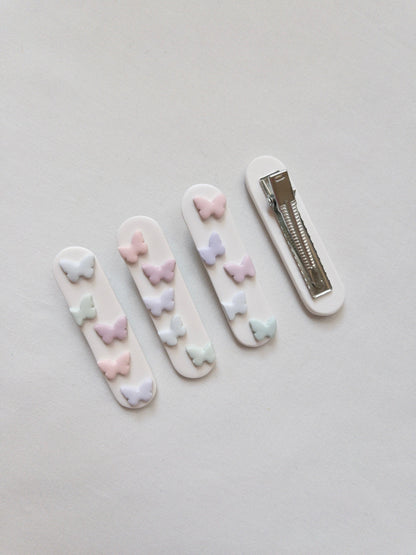 a range of white polymer clay hair clips with pastel butterfly accents on top with one upside down to show the stainless steel alligator hair clip on the back