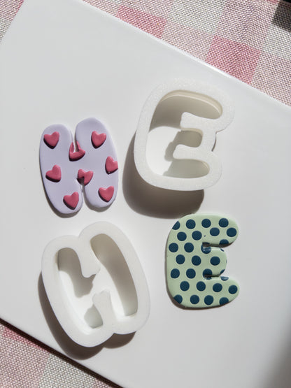 Four letter cutters in white silicone, shown with two decorated sample magnets: one lavender with pink hearts and one mint green with dark blue polka dots, on a white background with a pink checkered cloth edge.