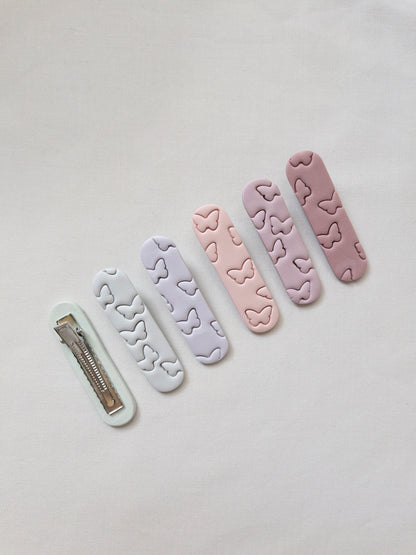 a range of pastel polymer clay butterfly printed hair clips with one upside down to show the stainless steel alligator clip on the bottom.